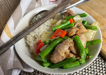 Ginger green bean and chicken stir fry served with brown rice in a white bowl with gray chopsticks and a spoon on the side.