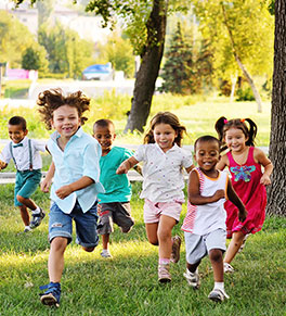A group of six, smiling children are running through a park.