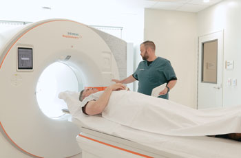 image of the new advanced photon counting computed tomography scanning machine at uci health irvine chao family comprehensive cancer center and ambulatory care building. a patient is inside the scanner while a tech in green scrubs sets up the equipment.