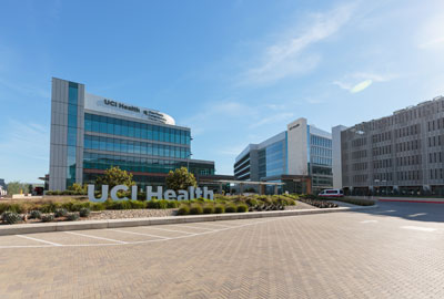 a view of the uci health irvine medical complex with the chao family comprehensive cancer center, acute care hospital and parking garage in the background behind a UCI Health sign