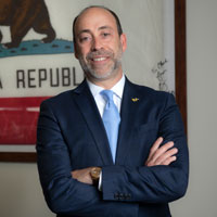 uci health ceo chad lefteris wearing a dark suit standing with arms crossed in his office in front of state of california flag