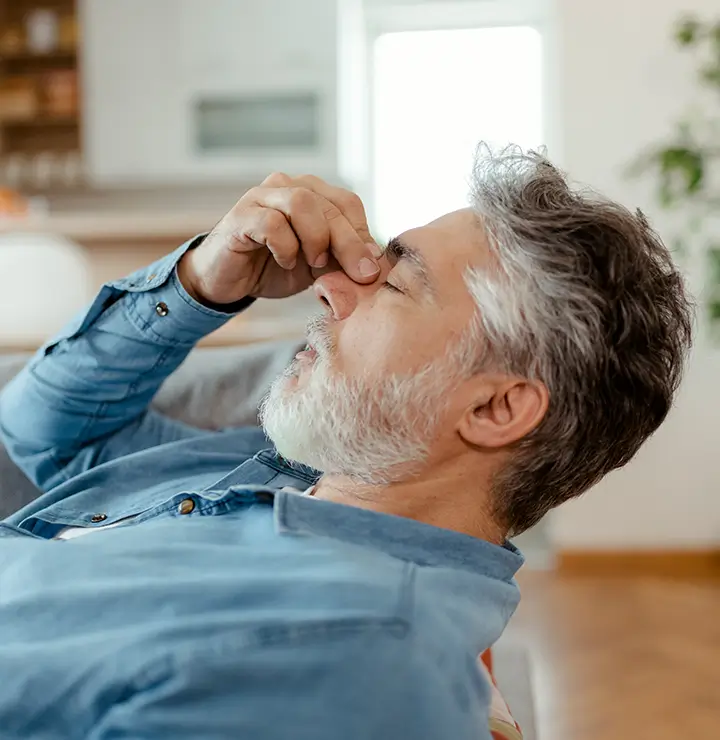 Tired man with gray hair and beard rubs his eyes on a couch with a blurred kitchen behind.