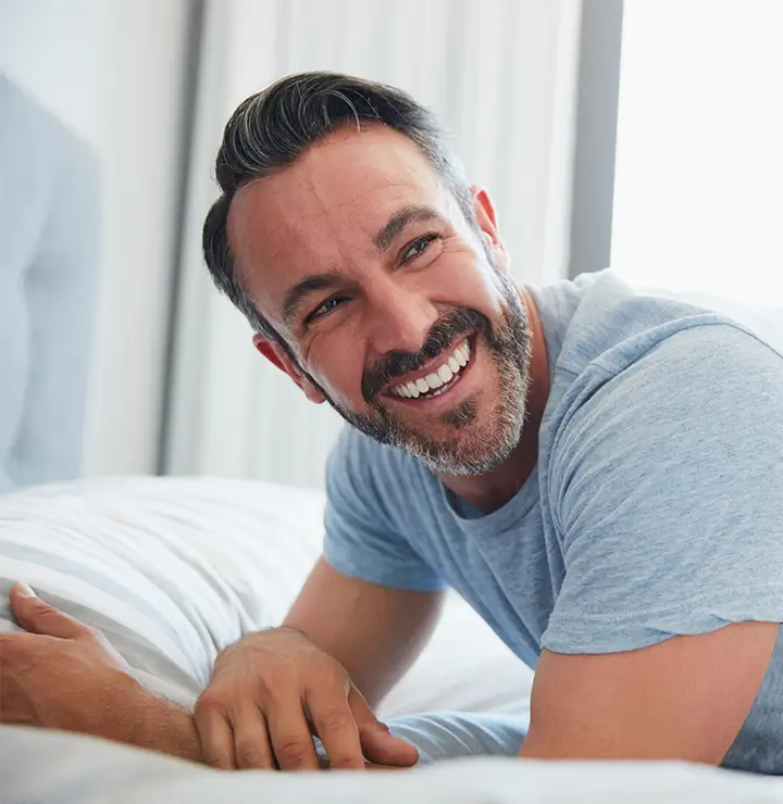A smiling man leans on a bed with striped sheets, wearing a blue t-shirt in a bright room.
